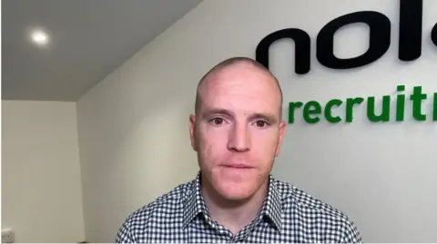 A man in a chequered white and black shirt is standing in front of a white wall with the black and green company logo in the background