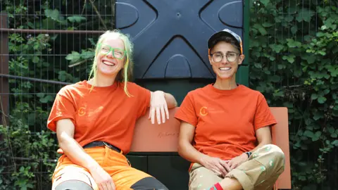 Steph Robinson is a white British woman with bleached blonde and grey hair. She wears green framed glasses, an orange t-shirt and orange working trousers with black patches over the knee and lower legs. ffin Jordao is Portuguese with short brown hair and light olive skin. They wear an orange and black peaked cap, the same orange work t-shirt as Steph and light beige working trousers. Both sit on a bench in front of metal fencing and greenery and smile at the camera.