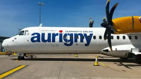 The side of a plane. It has Aurigny livery in blue colour text. It has black propellers on its wings and yellow wings. The sky is blue. It is taxiing on the grey tarmac and there are yellow cones. 
