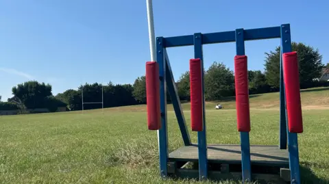 A low down image of a metal and wooden rugby scrum machine. It is painted blue and has red padding on the outside. Rugby posts are visible in the background as well as the pitch and a number of trees. 