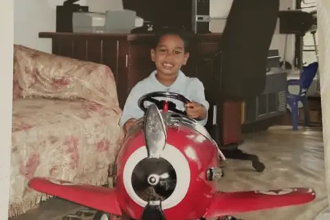 Family Shawn Seesahai as a young boy, sitting in a big red aeroplane toy while holding its steering wheel; he is sat in front of a desk with a computer and computer chair, and a sofa is to his left