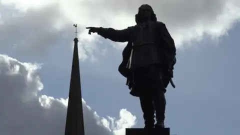 Somerset Film A statue, pictured in sillhouette and from below, showing a man in a long jacket pointing at the spire of an adjacent building. Blue skies and white fluffy clouds are visible in the background. 