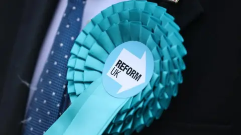 A light blue rosette pinned on a navy blue suit. The centre of the rosette has the Reform UK logo on it.
