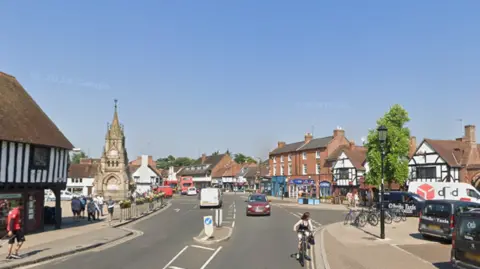 A street with shops, cars, and a clock tower in the centre, with people walking and cycling.
