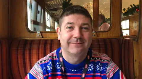 John Devine/BBC Chris O'Regan has short black hair and is wearing a blue, red and white Christmas jumper, with an icicle pattern on it. He has an NVR lanyard around his neck and is sitting in an old railway carriage