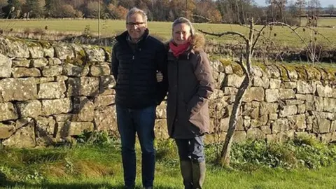 Charlotte and Charlie Bennett in front of a stone wall with fields and hills behind them