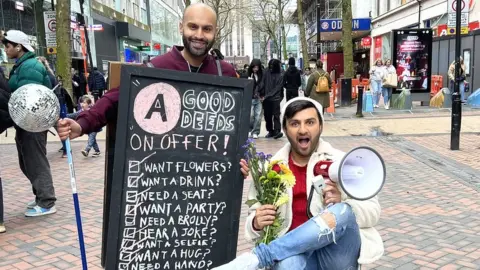 BBC Asian Network's Haroon Rashid - a bald man with dark beard - stands in a busy High Street wearing a chalk board with a list of good deeds on it. They are "Want Flowers? Want a Drink? Need a Seat? Want a Party? Need a Brolly? Hear a Joke? Want a Selfie? Want a Hug?" Shabaz Says sits next to him, holding flowers in one hand and a megaphone in the other. He's pulling a mock-surprised/shocked expression.