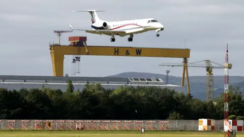 PA Media A plane carrying King Charles and Camilla, the Queen Consort, prepares to land at Belfast City Airport, with a Harland and Wolff shipyard crane in the background