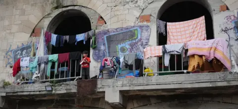 BBC A very young boy stands on a balcony with no barrier or railing at the derelict building, surrounded by washing that has been hung out to dry