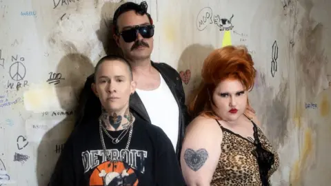 Cody Critchloe 2024 Promotional photo for the band Gossip, showing the trio posing against a concrete wall full of graffiti