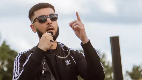 Ibrahim Ibrahim is holding a small mic to his mouth, and is wearing sunglasses - he's also in an Adidas black striped jacket