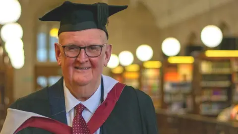 University of Bristol Paul Deal standing in a library wearing his graduation robes and looking at the camera