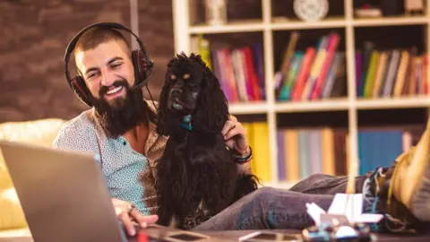 Getty Images A smiling man wears a headset with his feet up, cradling a dog while he taps on his laptop