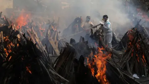 Getty Images Relatives stand next to the burning funeral pyres of those who died due to the coronavirus disease (COVID-19), at Ghazipur cremation ground in New Delhi.