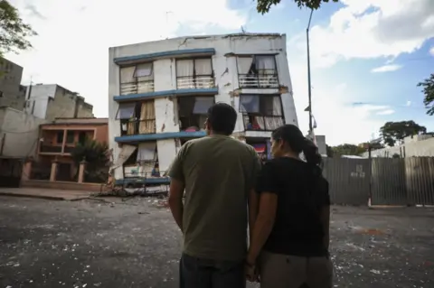 Clayton Conn Man and woman stood in front of a damaged building. What used to be a five-storey block has lost its bottom two levels entirely.