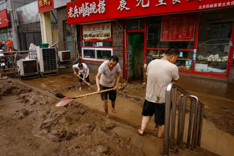 EPA Men shovel mud outside a store in Mentougou District, west of Beijing, China, 01 August 2023. Heavy rains brought by Typhoon Doksuri caused floods in northern China and left two dead and thousands being evacuated as Beijing experienced its heaviest rainfall of the year.