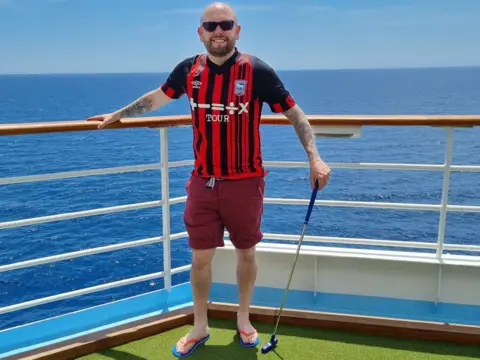 CHRIS PEACHEY Chris Peachey on a cruise ship, wearing a red and black Ipswich Town away kit