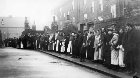 Newcastle Local Studies Library/Tyne Bridge A black and white picture of a line of people next to a brick wall and buildings