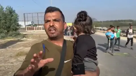 A picture of a man holding his toddler as he walks down Salah al-Din Road towards southern Gaza