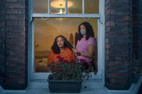 Julia Fullerton-Batten A woman combs a woman's hair next to a window, both wearing masks