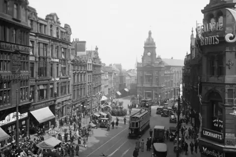 Newcastle Local Studies Library/Tyne Bridge Black and white of buildings, lots of people and trams on a street