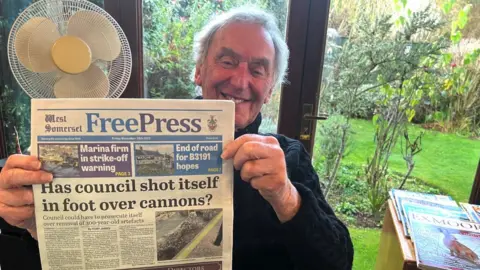 BBC Image of Tony James. He is holding a copy of the West Somerset Free Press and smiling.