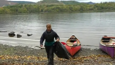 BBC Volunteer on island shore carrying a black refuse sack with canoes at waters edge