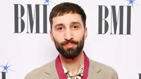 Getty Images Linden Jay at the BMI Awards. Linden, a young man, has short brown hair and a short dark beard. He has brown eyes and looks at the camera with a slight smile. He wears a beige suit over a cream coloured shirt with a maroon line pattern and a red scarf. He is pictured in front of a white back drop with the BMI logo printed on it.