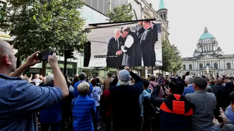 PA Media A crowd of people in Belfast city centre watch a large screen broadcasting the arrival of King Charles at St Anne's Cathedral