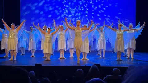 BBC A group of women in silver and white costumes dance on a stage