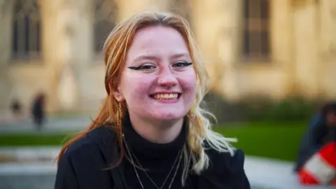 Blyth Lyes, a young woman with nose rings and eye make-up, smiling outside Gloucester Cathedral