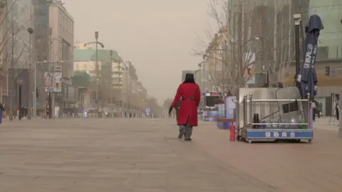 A guard in a red coat walks through an otherwise deserted shopping strip in Beijing