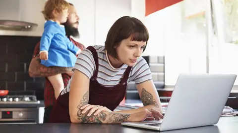 Getty Images Woman looking at laptop in kitchen with man and baby in background