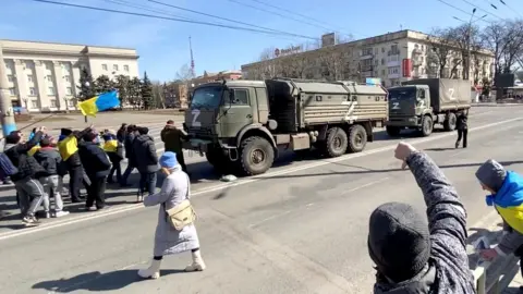 Reuters Demonstrators with Ukrainian flags chant "go home" to Russian military vehicles at a pro-Ukraine rally in Kherson, 20 March 2022