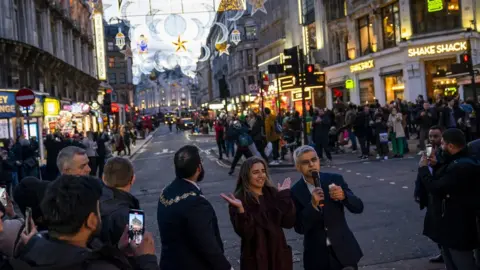 PA Media Mayor of London Sadiq Khan speaks into a microphone as people look on