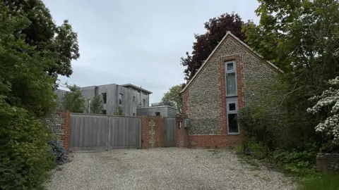 Martin Giles/BBC Arcady house, showing wooden structured house behind closed wooden gate and gravel driveway