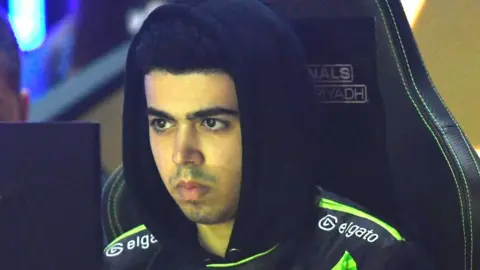 Mark Zakhary concentrates in a dimly lit arena as he competes in the FIFAe World Cup 2023. He's sat in a gaming chair, and he's wearing a black hoodie - with hood up - with lime green piping on the shoulders and a lime green slash across the chest. It says "elgato" in white on both shoulders. His face is tinged green from the monitor light.