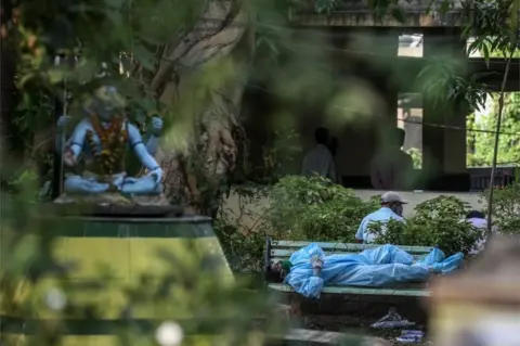 EPA A relative wearing personal protective equipment (PPE) rests on a bench from exhaustation before performing the last rites for COVID-19 victims during their funeral at a cremation ground in Vasai, on the outskirts of Mumbai, India, 13 April 2021