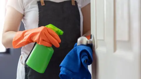 Getty Images Female hands in orange rubber gloves cleaning on touching surface of door, doorknob with blue microfiber cleaning cloth and green spray bottle - stock photo