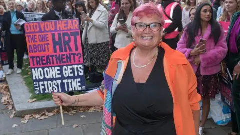 MP Carolyn Harris Carolyn Harris smiling wearing an orange jacket with pink hair and dark glasses at the front of a crowd holding a placard about HRT