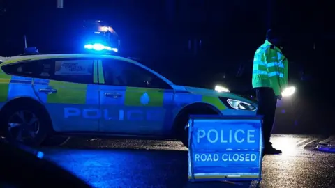 Dan Jessup A police officer and police car with a road closure sign at night