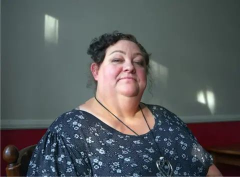 BBC Helen Murphy, wearing a blue patterned dress, sitting at a wooden table. There are streaks of light coming in from the left of the frame which light up parts of the wall behind her.