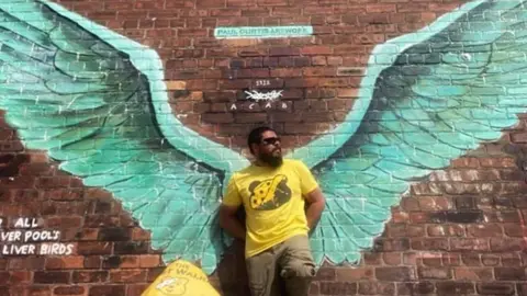 Chris Howard Chris Howard in front of a mural of angel wings in Liverpool