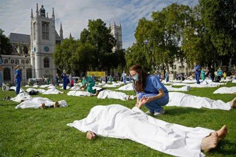 Neil Hall/EPA Protesters lie on the ground pretending to be dead with white sheets over them, with doctors looking on