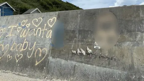 Steve Hobart Photography Graffiti on sea wall at Cromer, including 'I want a baby' and hearts in spray paint, with some spraypaint on part of the Banksy mural