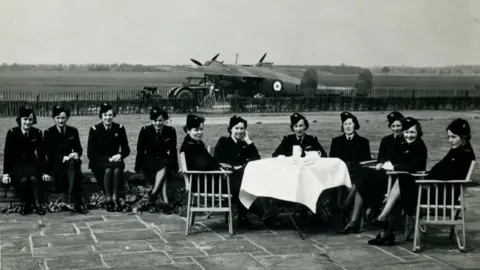 P Gower Photos / Air Transport Auxiliary Museum A black and white photo of an airfield where 10 women in uniform and hats are sitting around a table in front of an RAF De Haviland Flamingo aircraft