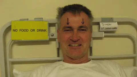 Simon St Leger-Harris Man in hospital bed with two arrows drawn on above his eyes