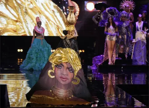 EPA A woman walks down a catwalk wearing a black and metallic outfit, featuring a large pot on her head and an embellished image of a woman's face on her dress.