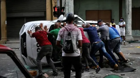 YAMIL LAGE/AFP People push an overturned car in the street in the framework of a demonstration against Cuban President Miguel Diaz-Canel in Havana, on July 11, 2021.