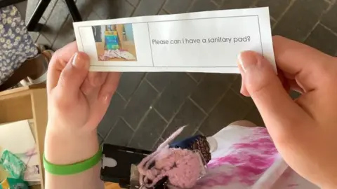 Portland College Photo shows student holding a slip that says 'please can I have a sanitary pad?' next to a photo of some pads. The student is handing it in to reception.